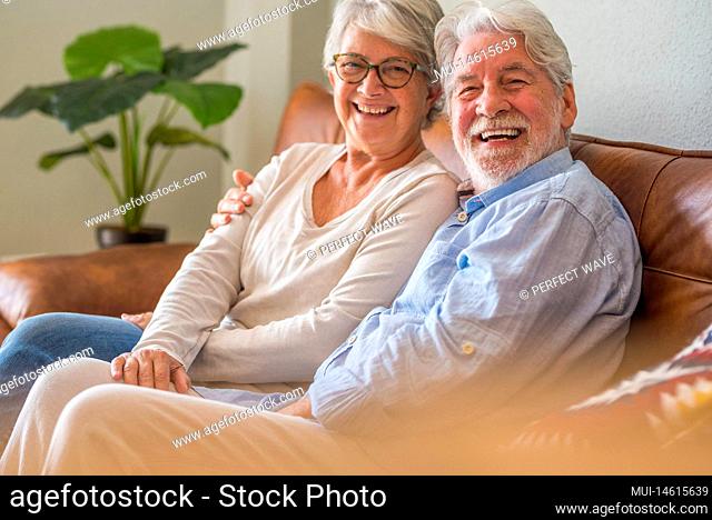 Portrait of cheerful senior couple embracing while sitting on sofa and smiling. Elderly happy couple relaxing and posing in front of camera sitting in living...