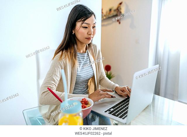Young woman eating breakfast, while working on laptop