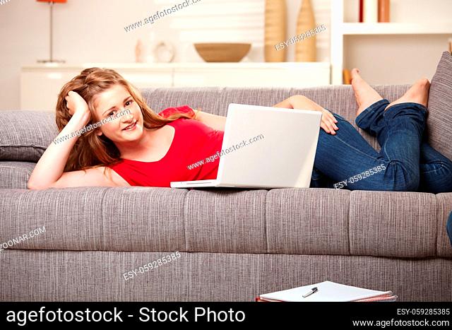 Smiling teen girl with laptop computer resting on sofa at home browsing internet looking at camera