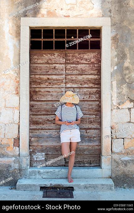 Beautiful young female tourist woman standing in front of vinatage wooden window and textured stone wall at old Mediterranean town, smiling, holding