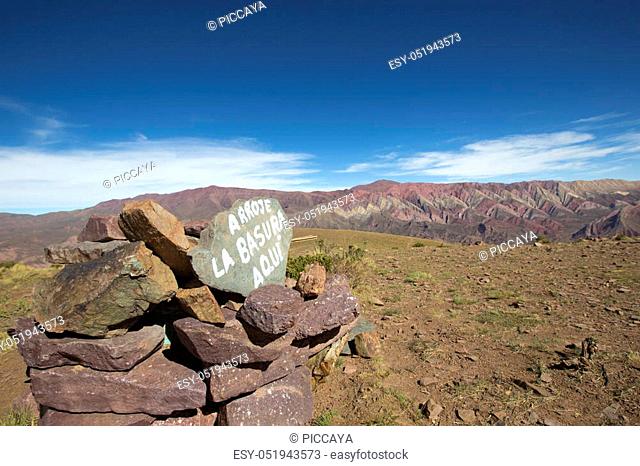 Pile of stones and sign in Spanish saying ""Garbage is Here"" with the Mountain of fourteen colors, Quebrada de Humahuaca against a blue sky in the background