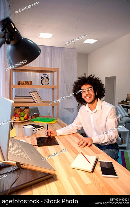Happy hipster freelance man working at table and smiling for camera. Handsome man in glasses sitting at table and using editing board at home