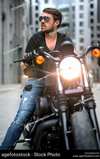 Bearded man sits on the black motorbike on the skyscraper background. Headlamp switched on. He wears a blue ripped jeans, a black T-shirt