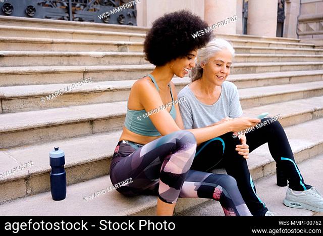 Smiling daughter sharing smart phone with mother sitting on steps