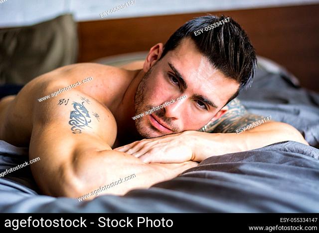 Shirtless sexy muscular male model lying alone on his bed in his bedroom, looking at camera with a seductive attitude