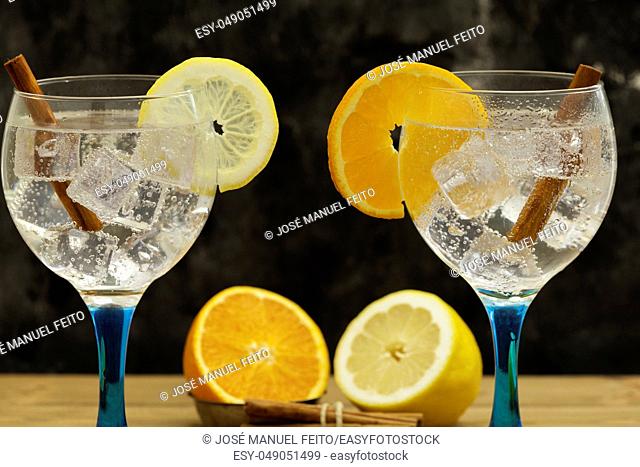 Two gin tonics on blue glass with lemon slice and orange slice, lemon and orange fruit and cinnamon sticks on wooden table and dark backdrop midplane
