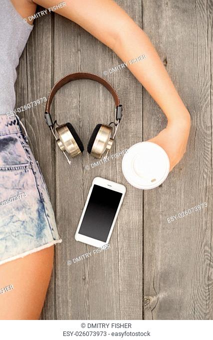 Beautiful young woman lying on the wooden floor with music headphones, smartphone and a take away coffee cup; view from above