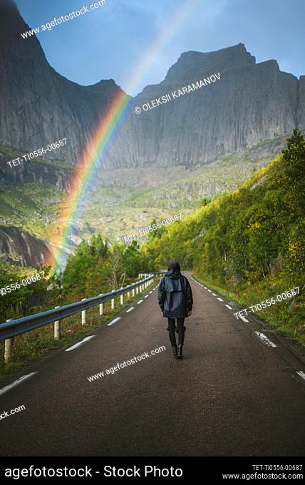 Norway, Lofoten Islands, Man walking down road with mountains and rainbow in background