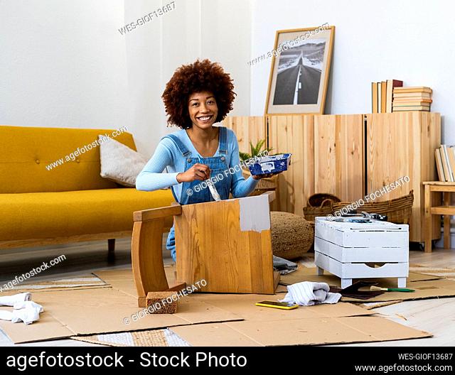 Happy young woman with paint tray painting old chair in living room