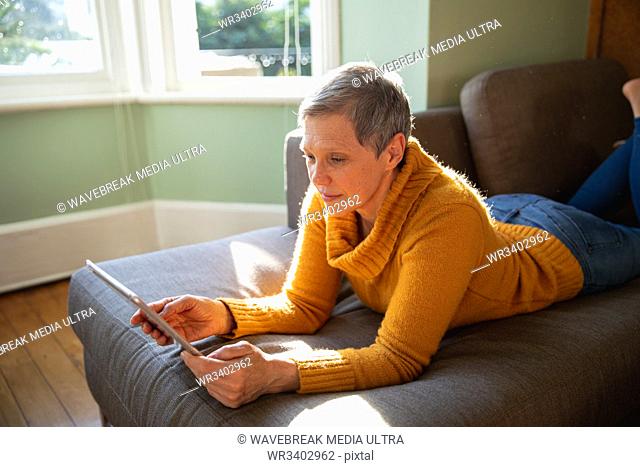 Side view close up of a mature Caucasian woman with short grey hair lying on her front on a sofa in her living room, leaning on her elbows and using a tablet...