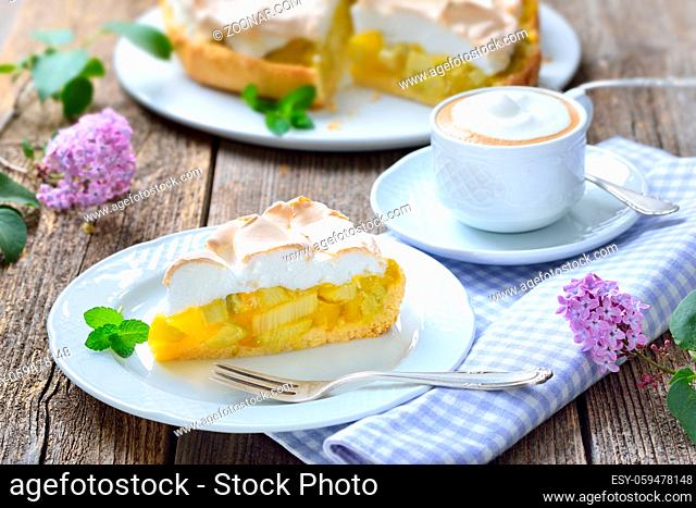 Leckerer Rhabarberkuchen mit Baiserhaube und Cappuccino auf einem Holztisch mit fruehlingshafter mit Fliederdeko - Delicious rhubarb cake with springlike lilac...