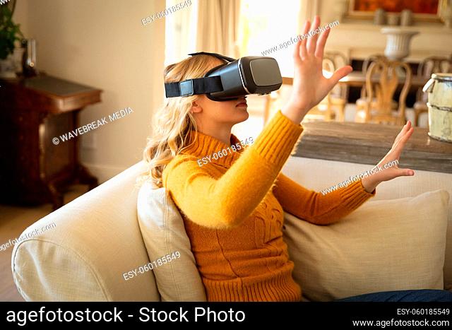 Caucasian woman sitting on couch in luxury living room wearing vr headset, with hands raised