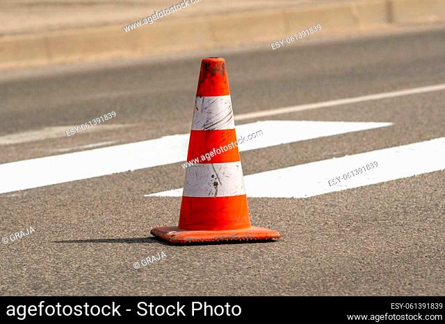 Traffic cone with orange and white stripes standing on street on gray asphalt during road construction works. Just painted white street lines on pedestrian...