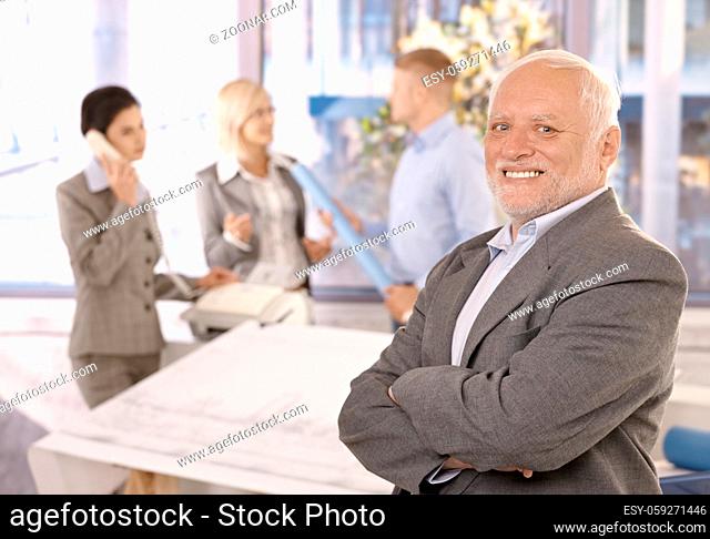 Proud smiling senior businessman standing with arms crossed, smiling, with team working in background