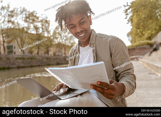 Self-employment. Smiling african american guy looking at papers in hand working on laptop sitting near water outdoors on warm day