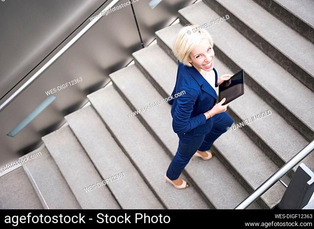 Portrait of smiling blond businesswoman climbing stairs with tablet