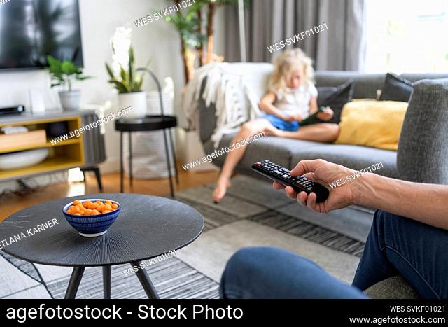 Hand of man holding remote control with daughter in background on sofa