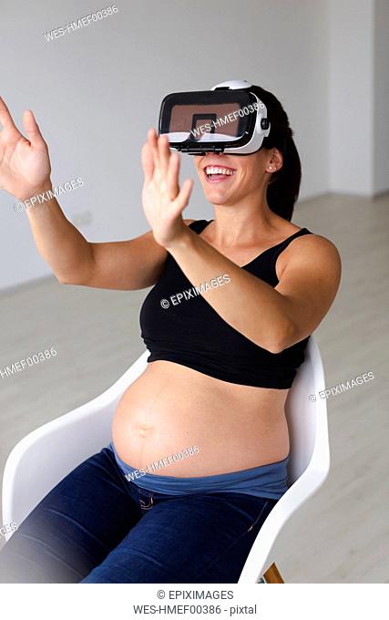 Young pregnant woman sitting in an armchair and watching and feeling her baby with the help of virtual reality glasses