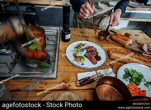 Male chef seasoning sauce on tomahawk steak in plate while standing at kitchen