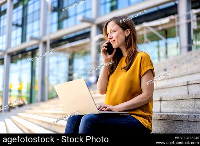 Smiling businesswoman sitting with laptop talking on mobile phone