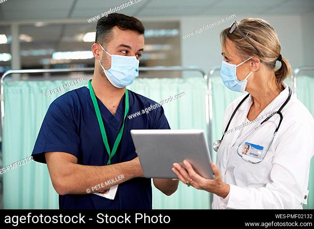 Male nurse with protective face mask looking at doctor holding tablet PC in medical room