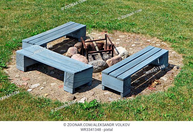 The place for a barbecue on a green grass of a garden lawn. Wooden handmade benches are painted with gray paint. The brazier is made of granite stones