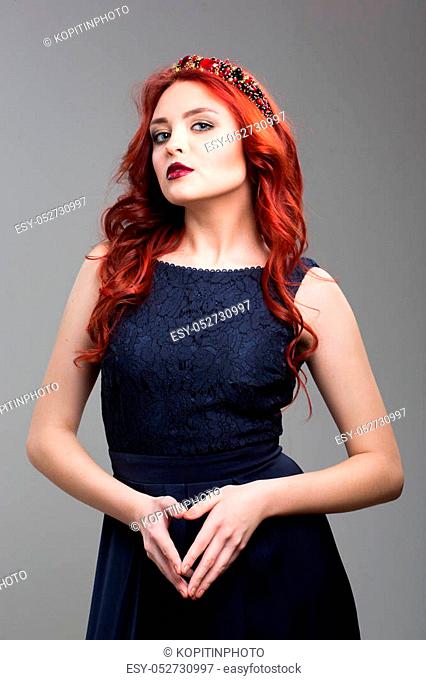 Beautiful red-haired fashion model posing in evening dress and in the diadem over dark background. female gestures of seduction. body language