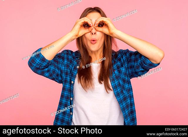 Wow, unbelievable. Portrait of surprised pretty girl in checkered shirt looking through fingers in binoculars gesture, amazed and shocked by seen