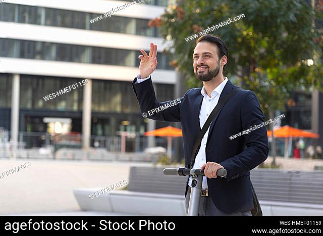 Smiling businessman waving on electric scooter in city