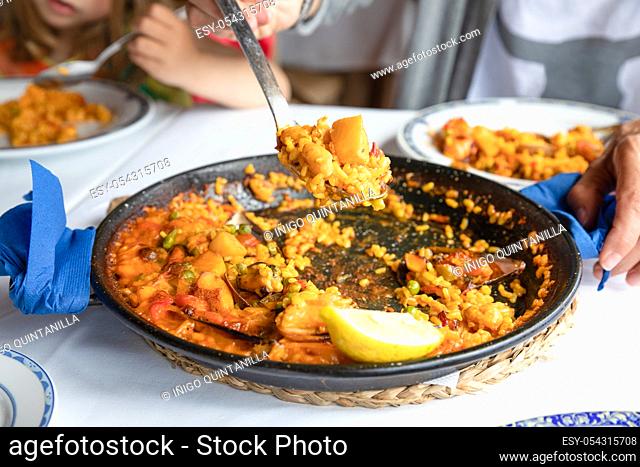 serving typical Spanish paella with seafood (squid) and seashell (mussel, clam), rice and lemon with big spoon on white tablecloth of restaurant