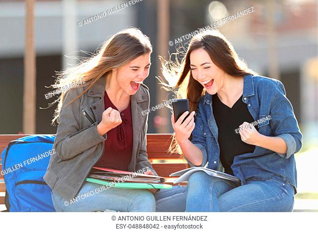 Excited students checking grades on smart phone sitting on a bench in a park