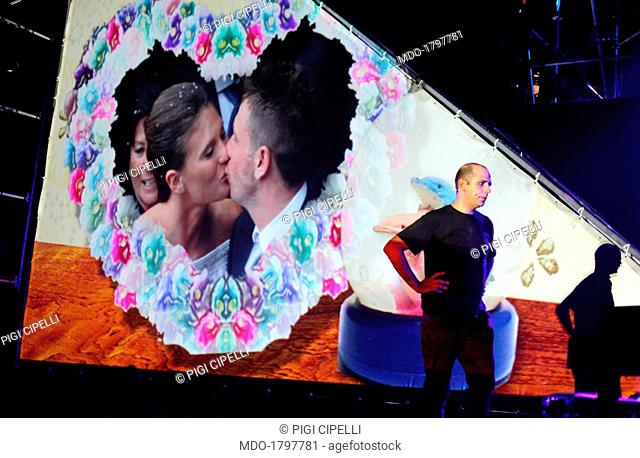 Checco Zalone on stage in front of a widescreen projecting an image of the wedding of Antonio Cassano and Carolina Marcialis