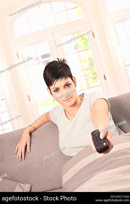 Young woman is watching TV at home in the living room with a remote controller in hand