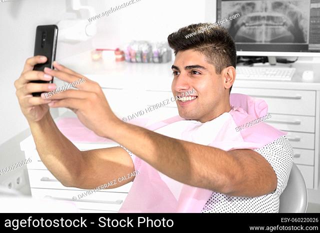 Happy patient taking selfie at dental clinic. Patient is holding smartphone and showing his healthy smile. High quality photo
