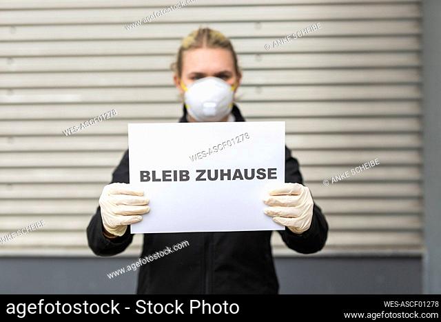 Teenage girl wearing protective mask and gloves holding sign with request 'Bleib Zuhause'