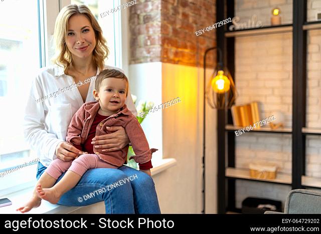 Blonde attractive woman with infant baby sitting on windowsill and playing together expressing happiness