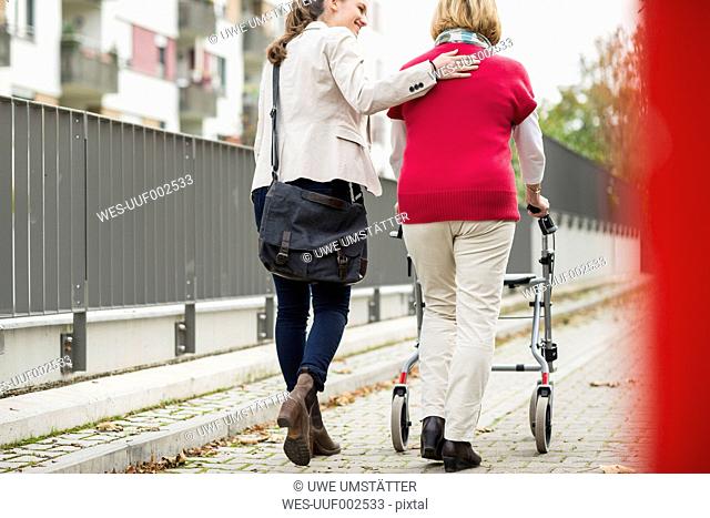 Adult granddaughter assisting her grandmother walking with wheeled walker, back view