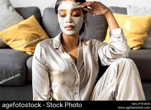 Serious woman with beauty facial mask sitting in living room at home