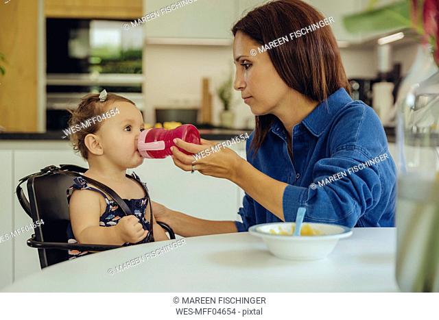 Mother helping baby daughter drinking water from bottle in kitchen