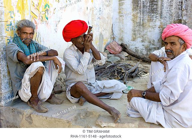 Indian man in traditional clothing smokes clay pipe while sitting with friends in Narlai village in Rajasthan, Northern India