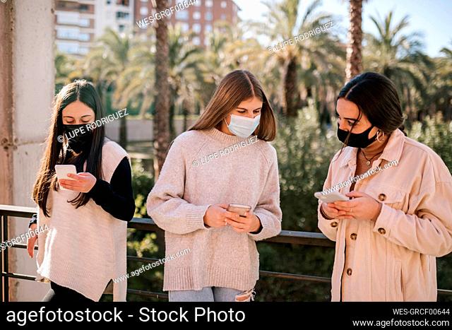 Teenage friends wearing face masks using mobile phones while standing in park