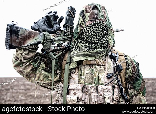 Army marksman, airsoft player in camouflage uniform and load carrier, masking cape on head, armed service rifle with optical sight, hiding face with shemagh