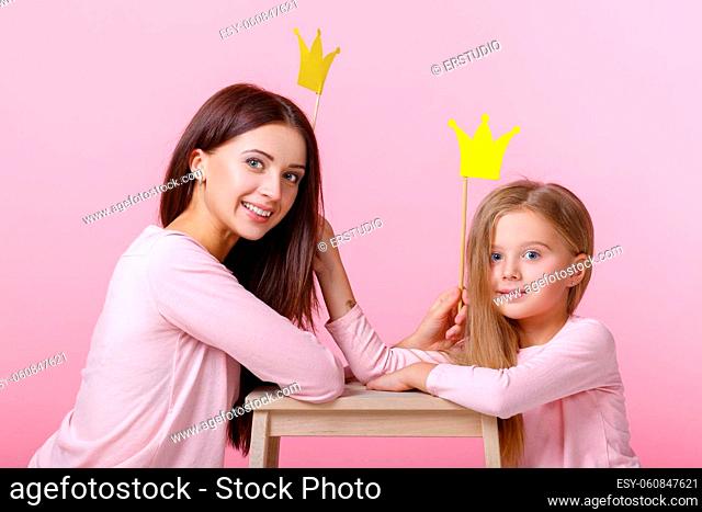 young mother and daughter having fun together and holding paper yellow crown on pink background
