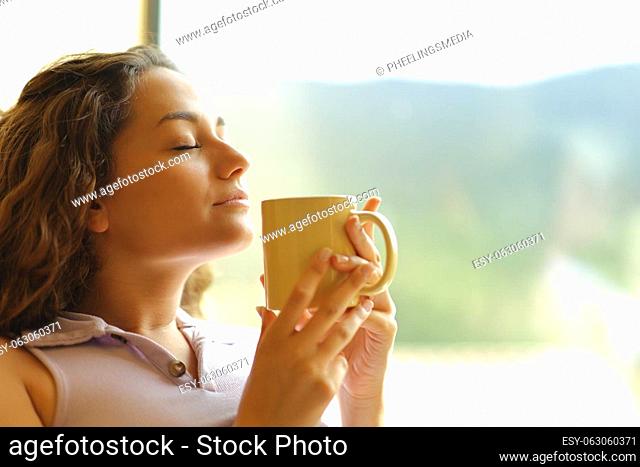 Happy woman smelling coffee relaxing beside a window at home with closed eyes