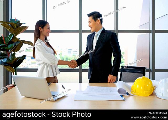 Asian project owner in suit and his Business partners shaking hands after checking the construction drawing. Morning work atmosphere in a modern office