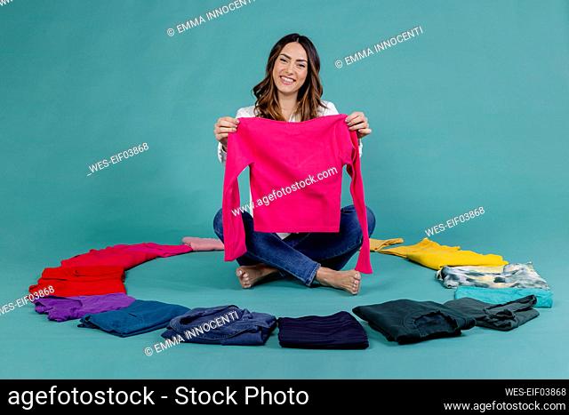 Smiling woman holding pink t-shirt sitting cross-legged amidst clothes against blue background