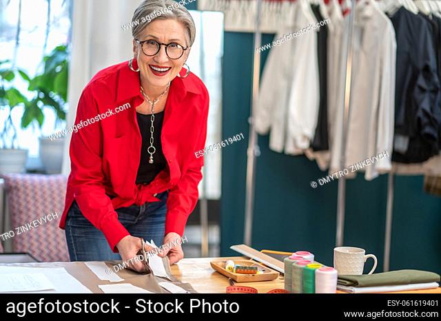 Lucky day. Happy middle aged woman in glasses looking confidently at camera standing near table cutting out pattern in atelier