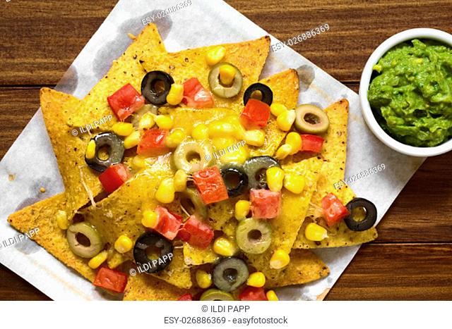 Baked nachos with cheese, green and black olives, tomato and corn, with guacamole on the side, photographed overhead with natural light (Selective Focus