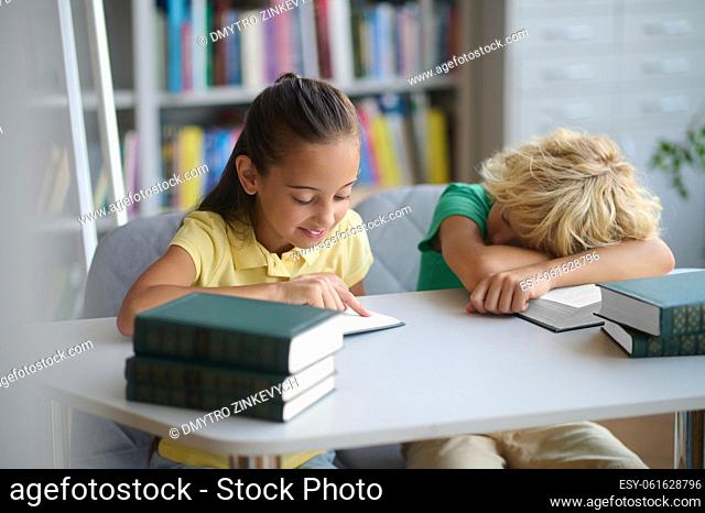 Smiling focused schoolgirl seated at the table reading while a schoolboy dozing on an open textbook