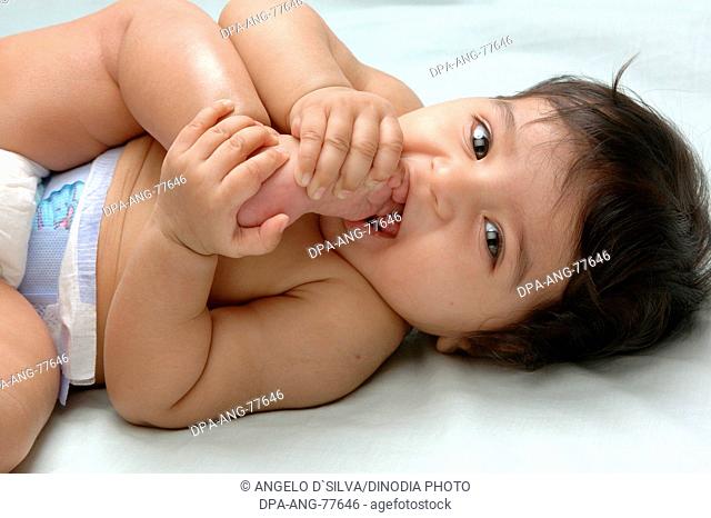Young baby Indian girl in diapers ; 4 months old ; Holding her feet putting her toe in the mouth and smiling ; Model Release 468